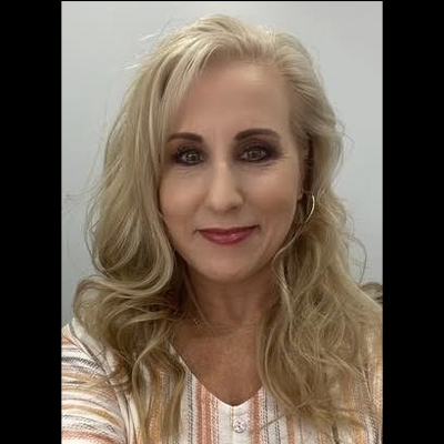 A woman with wavy blonde hair is wearing a light-colored striped top, posing in a neutral-toned environment.