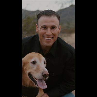 A person in a black shirt poses with a golden retriever against a backdrop of mountains and desert vegetation.