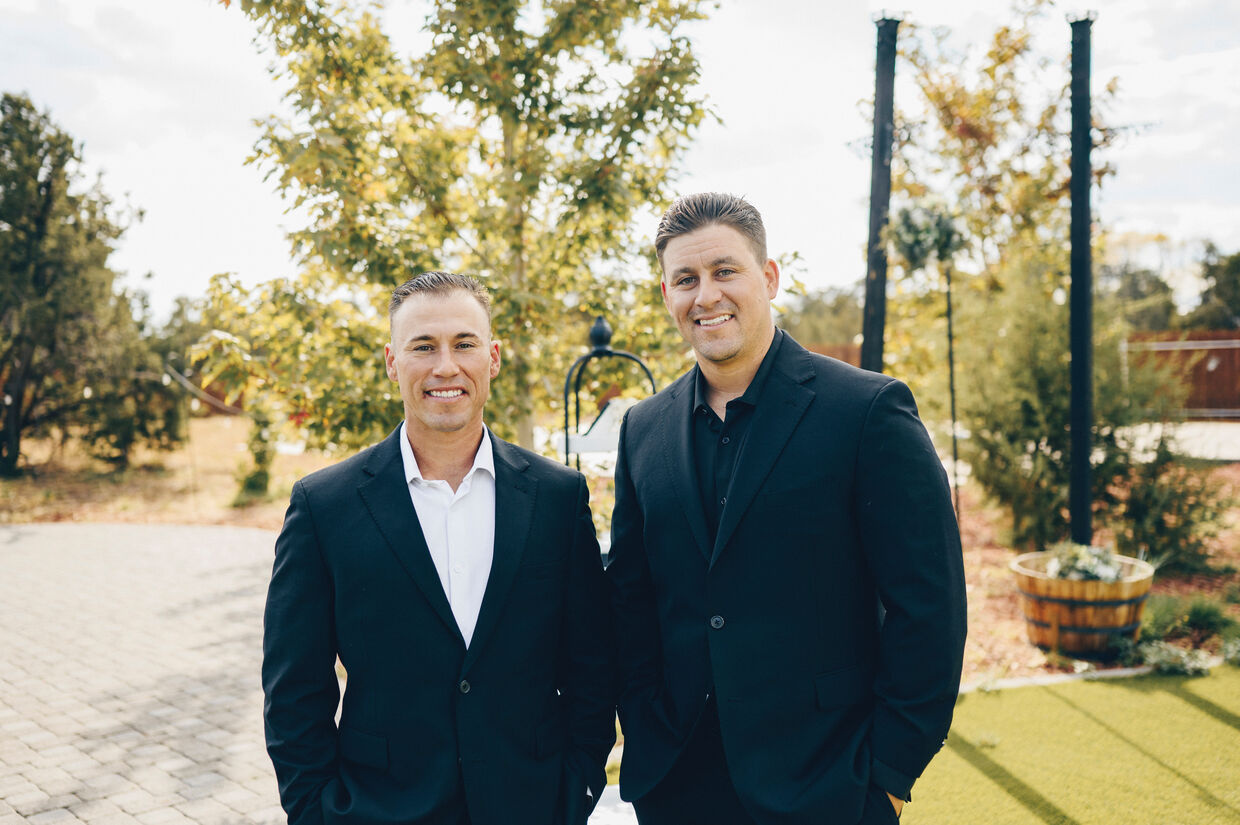 Two men in suits stand together outdoors, surrounded by trees and greenery, exuding a stylish and professional vibe.